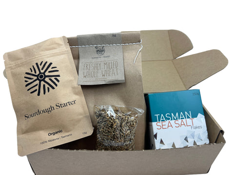 Box containing a bag of sourdough starter, a packet of whole wheat flour, a bag of seeds, and a container of Tasman Sea Salt flakes.